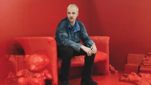Glenn Martins sitting on a red sculptural bench against a red background, wearing a denim jacket and black pants.