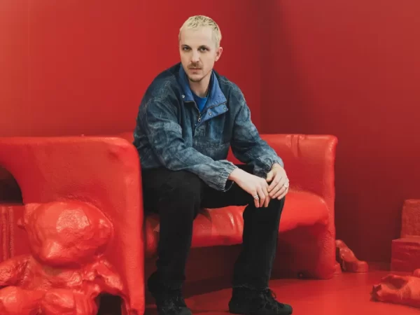 Glenn Martins sitting on a red sculptural bench against a red background, wearing a denim jacket and black pants.